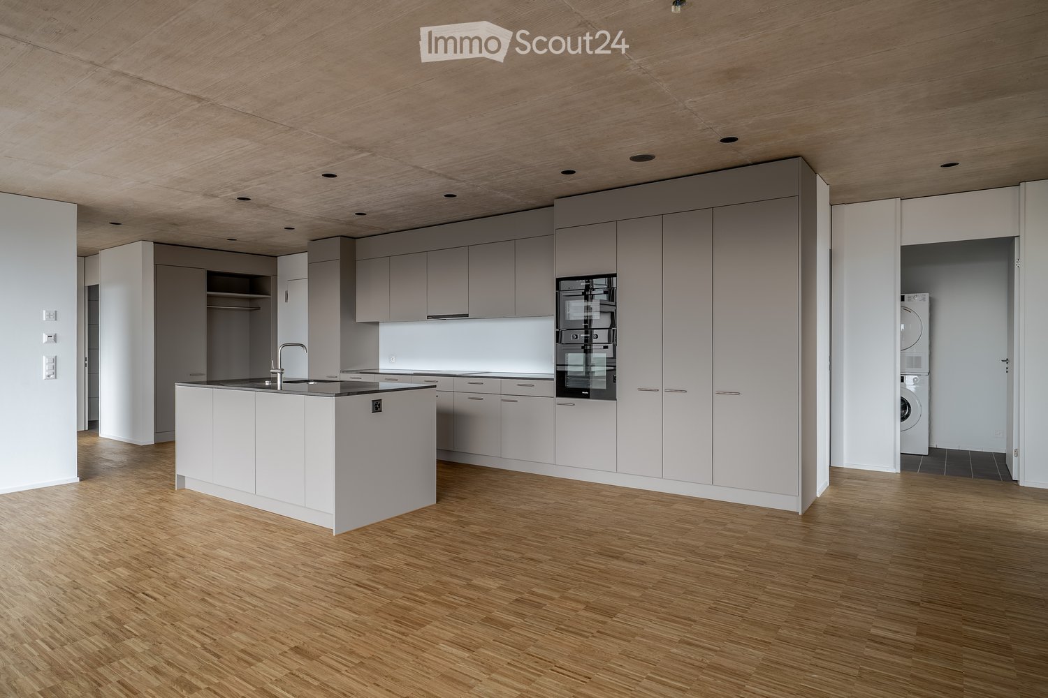 modern kitchen with white cabinetry, hardwood floors, central island with sink, integrated appliances including oven, dishwasher, and refrigerator, and a separate laundry room with washing machine