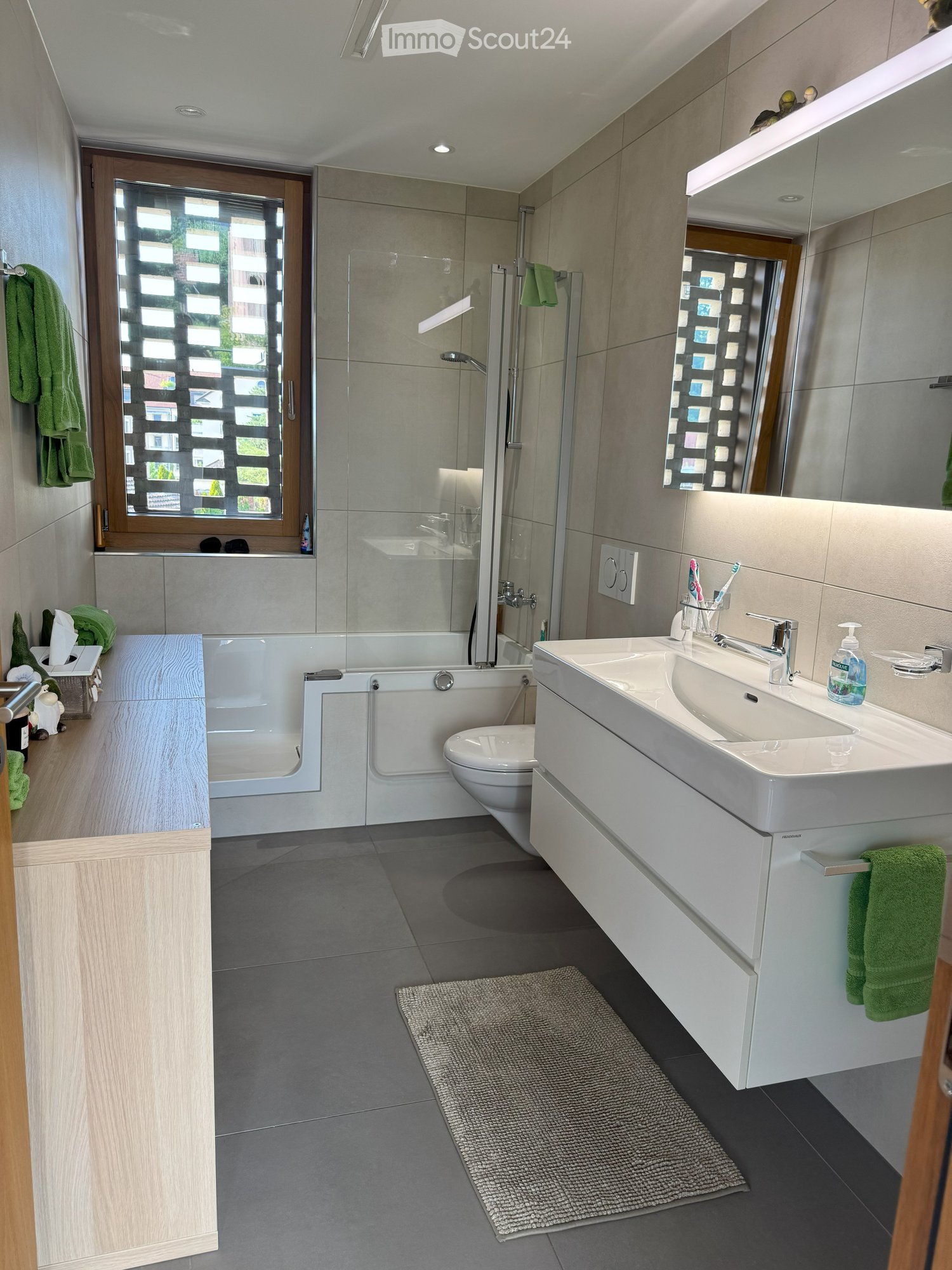 Modern bathroom with gray tiles, white bathtub, sink with mirror, wooden cabinet, toilet, and window with green curtains.