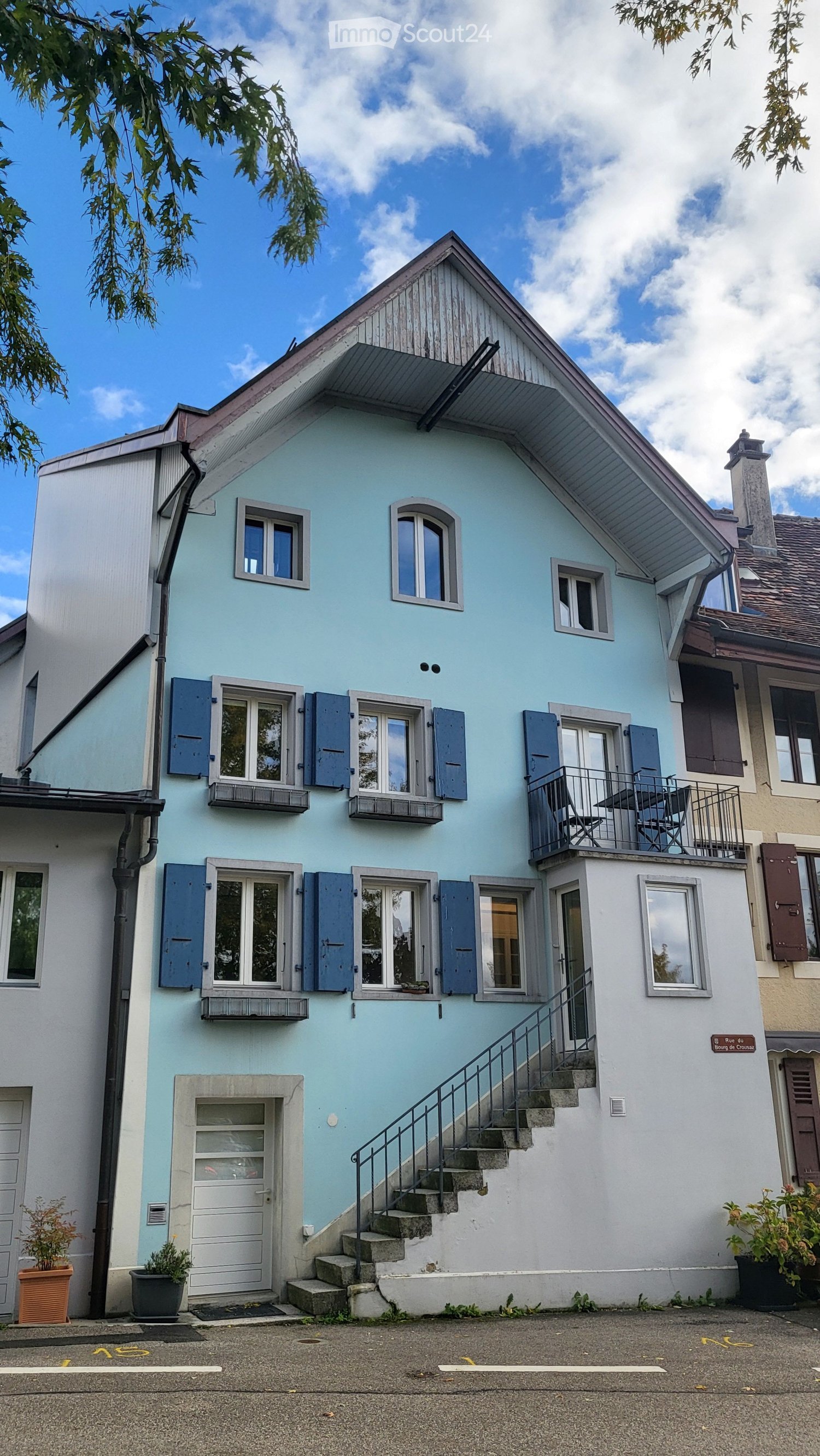 Two story house, blue exterior, white trim, multiple windows, balcony on the second floor, entrance with white door, outdoor seating on balcony