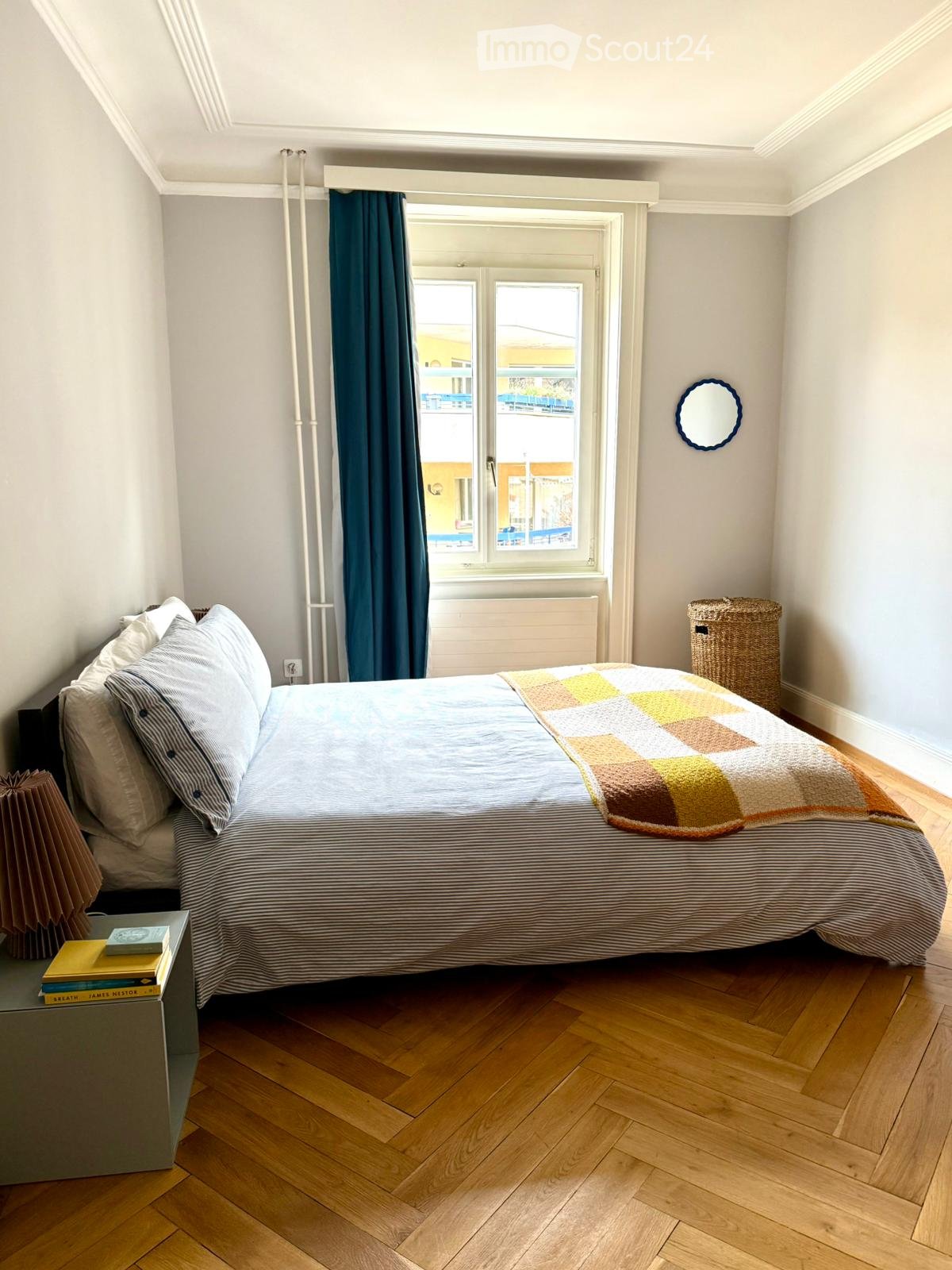 double bed with pillows, wooden floor, white walls, large window with curtains, bedside table with books, wicker basket, mirror