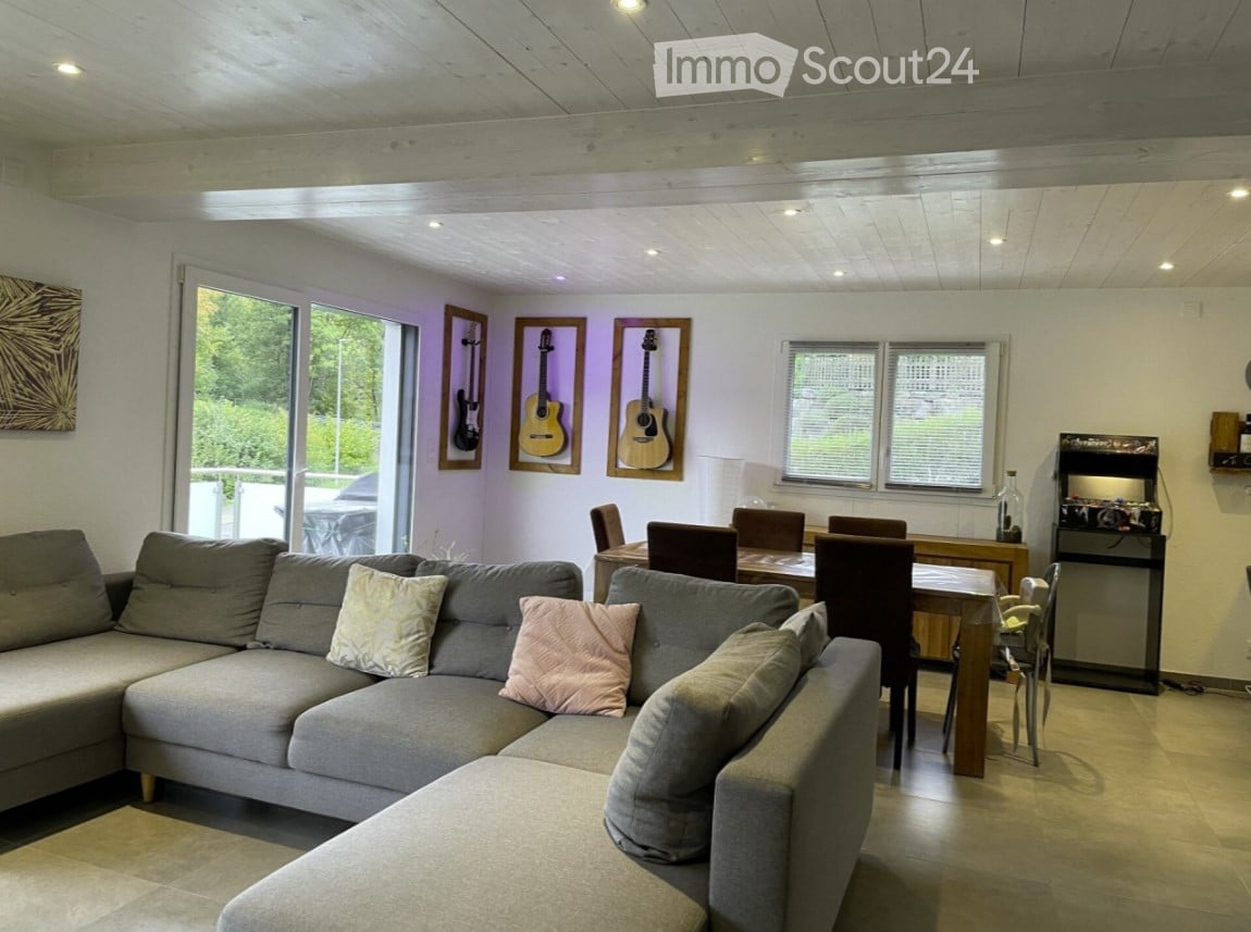 a living room with a gray L-shaped sofa, wooden table and chairs, guitars on the wall, and large glass windows overlooking trees outside