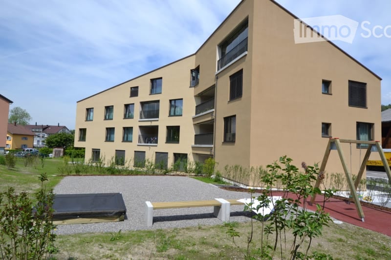 3-story apartment building, beige exterior, numerous windows, balconies, playground, bench, swing, paved area