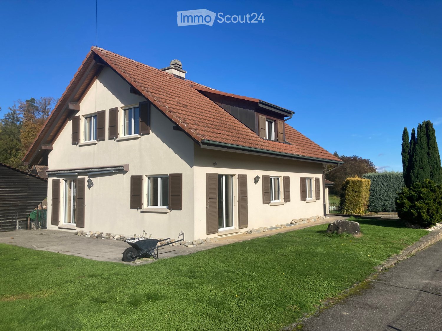 2-story house, brown roof, white facade, brown shutters, paved driveway, lawn, wheelbarrow, garden