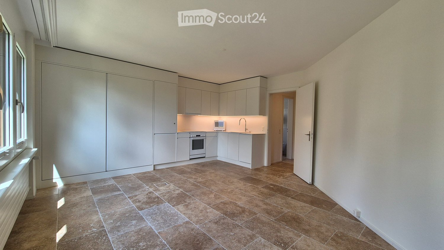 An empty kitchen with modern appliances and a bright atmosphere. It features a sleek white cabinetry, tiled flooring, and a modern kitchen island with a sink, faucet, and microwave. There is a white oven under the countertop. The room is well-lit by natur