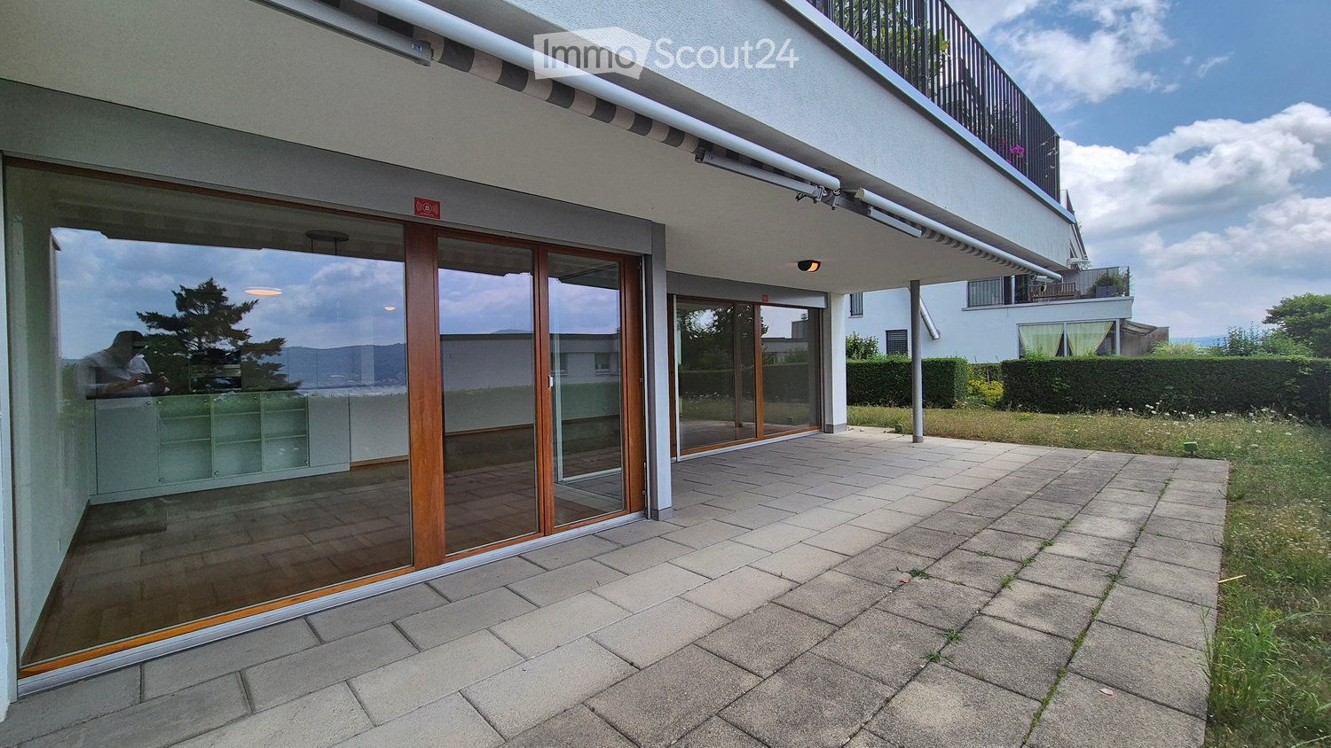 Modern apartment with large windows, balcony, paved outdoor area, reflecting trees and sky, well-maintained garden with shrubs, distant mountains