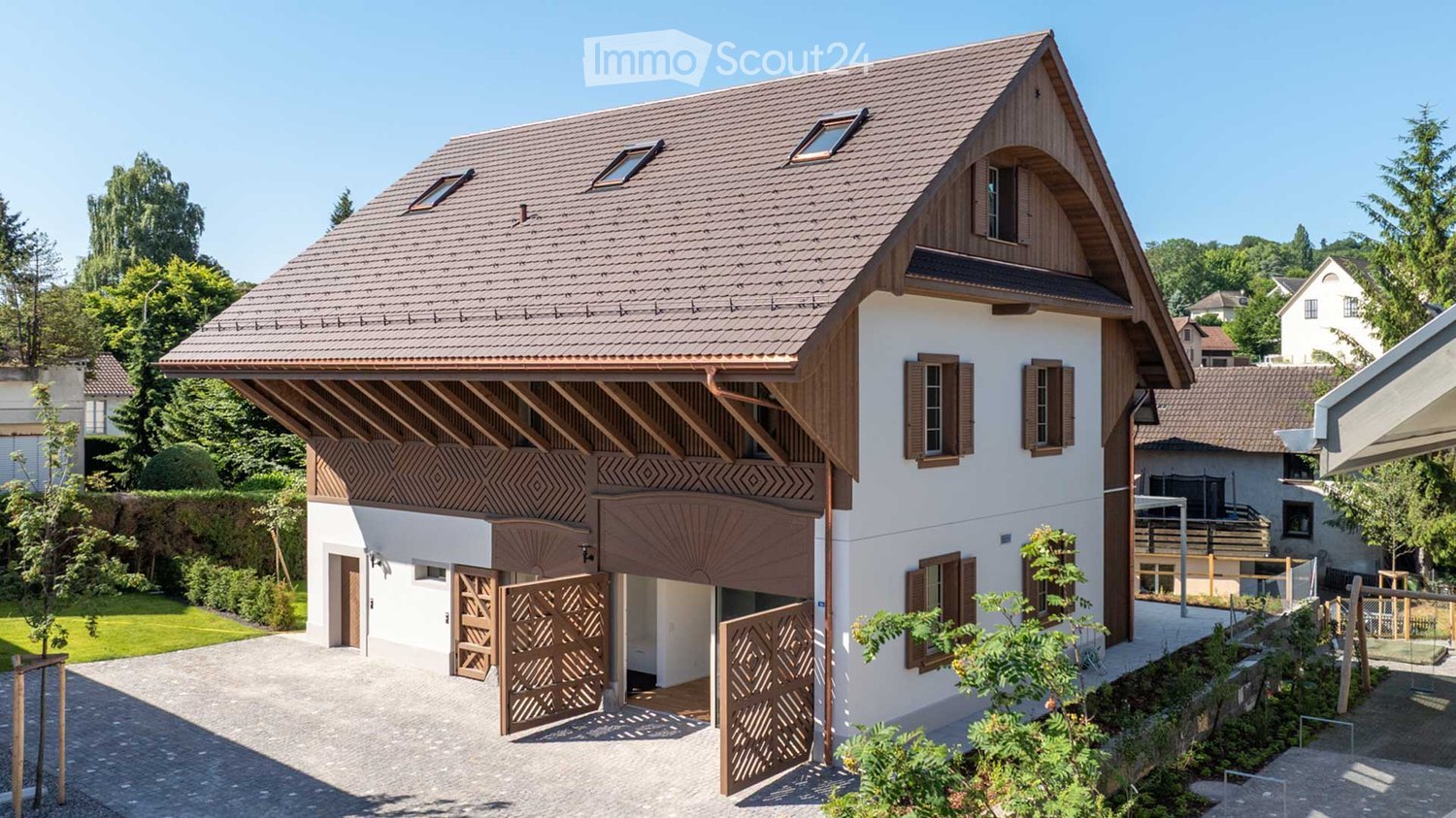 Two-story wooden house with a tiled roof, decorative wooden beams, and a garage with ornate wooden doors. The house is surrounded by lush greenery and trees.
