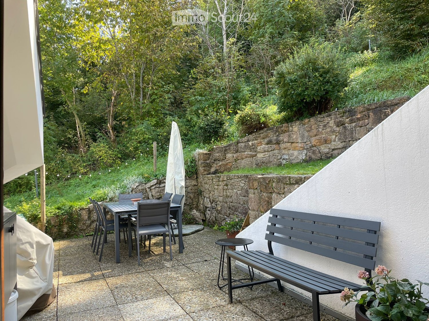 Patio with table and chairs, covered by an umbrella, next to a bench and potted plants, a stone wall is in the background
