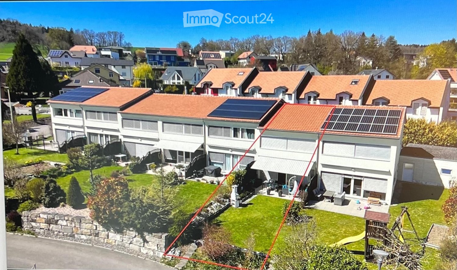 Large apartment building with solar panels on the roof, multiple levels, fenced garden, playground, pathway, several neighboring houses in the background.