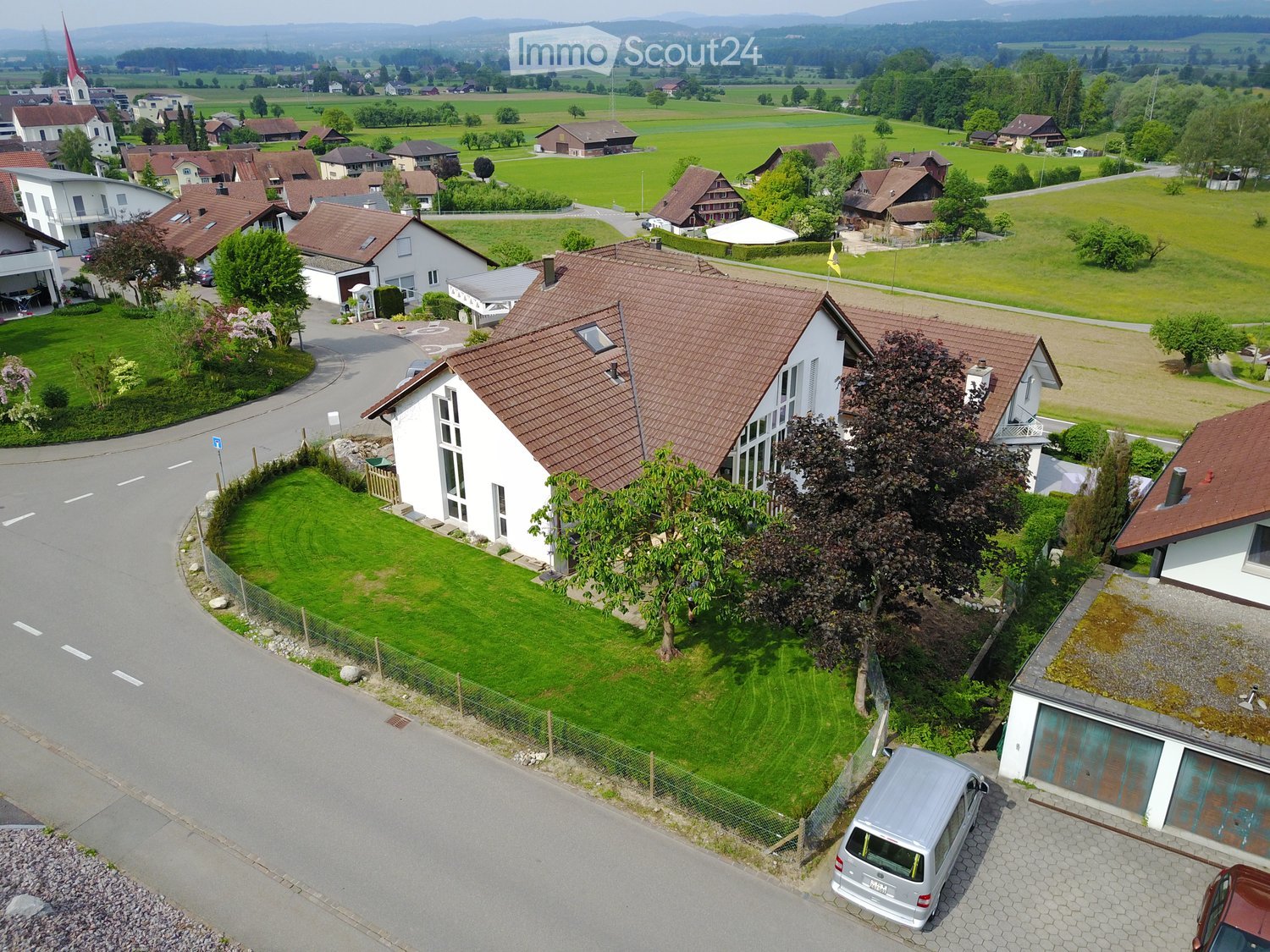 a large single-family house with brown roof and white walls, surrounded by a fence, located in a village with well-manicured lawns and green fields, a white van parked in the driveway, and an aerial view of the surrounding countryside with a church tower