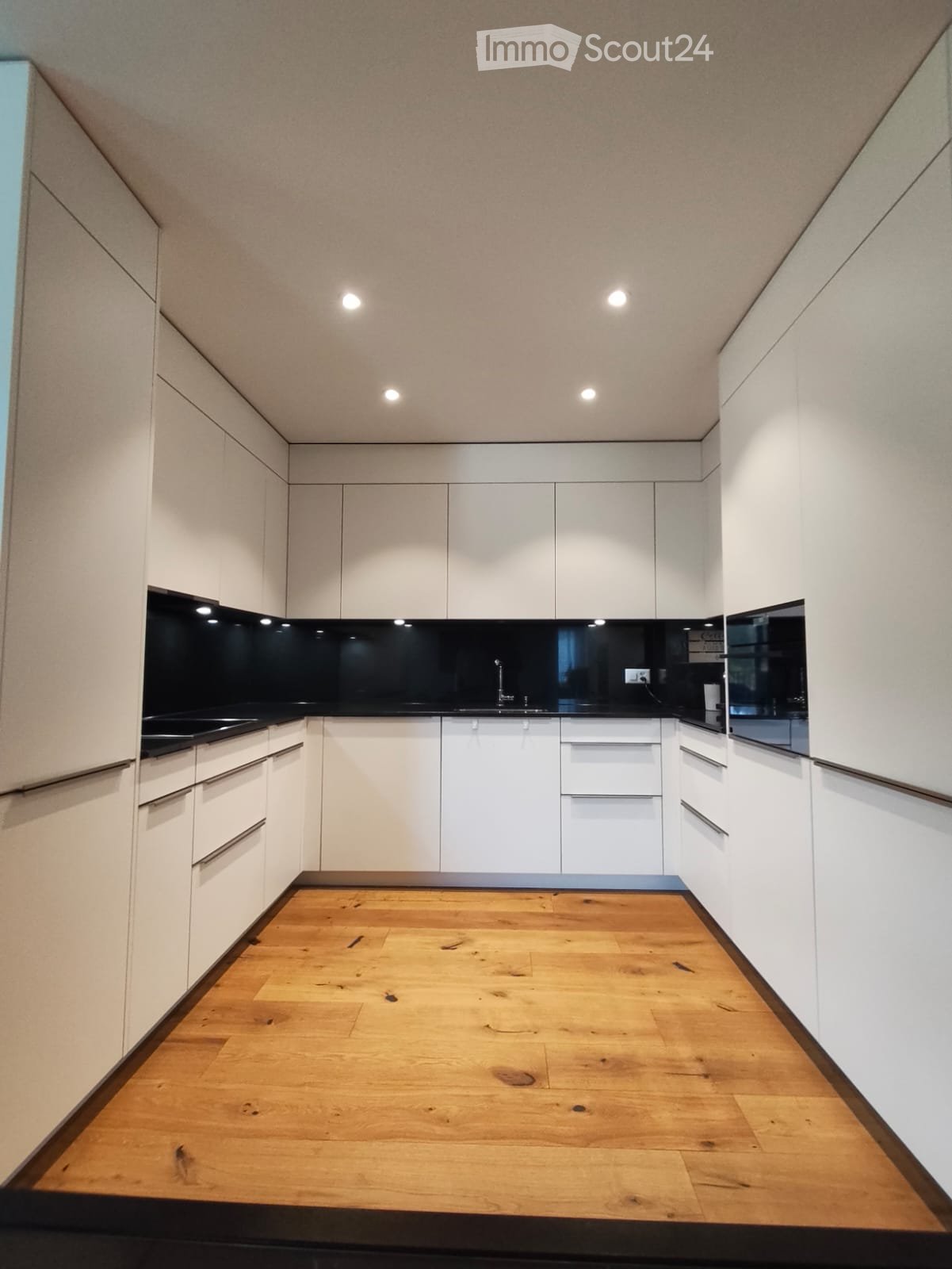 Modern minimalist kitchen, white cabinetry, wooden floor, black countertops, recessed lighting, integrated appliances