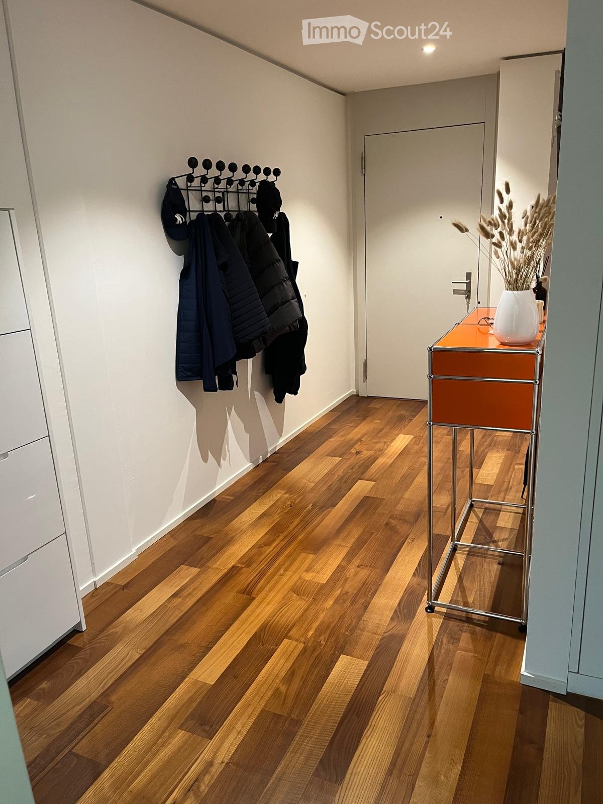 Entryway with white walls, hardwood floors, white cabinet with drawers, a coat rack on the wall, a white vase with dried flowers, an orange table with a metal stand