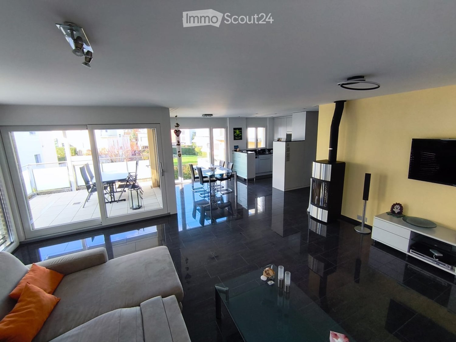 large living room with glass sliding doors leading to balcony, modern black tiled floor, orange couch with cushions, fireplace, TV, speakers, round table, modern kitchen with a dining table