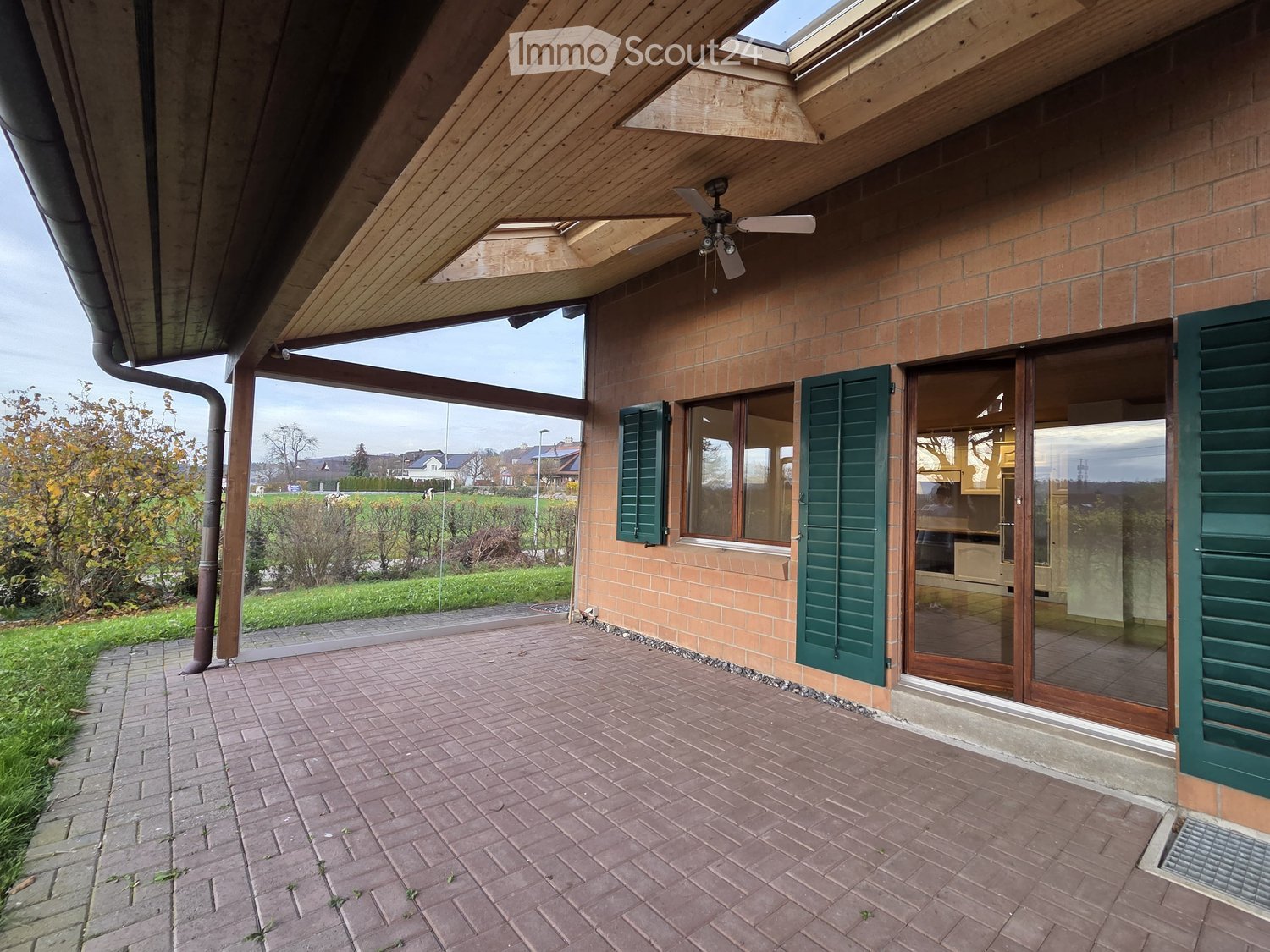 A brick house with a ceiling fan and a brick patio, two glass doors with green shutters and a garden view.