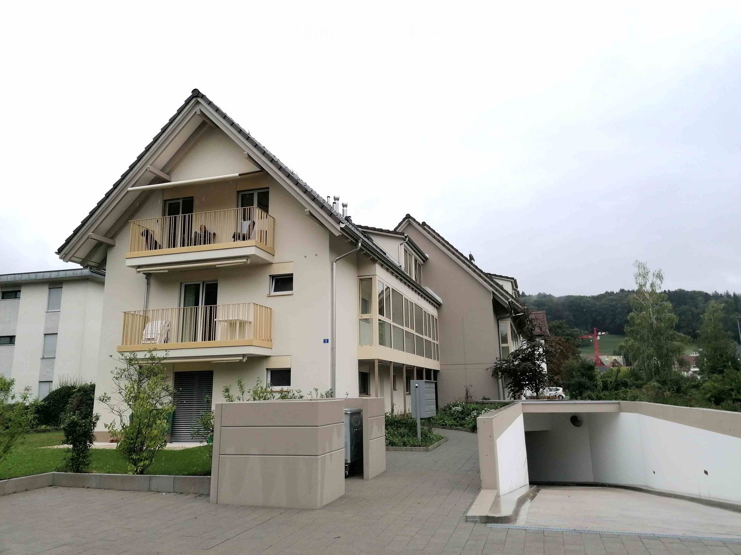 White apartment with beige balconies, concrete walkway, trash bin, large front yard, trees, bushes