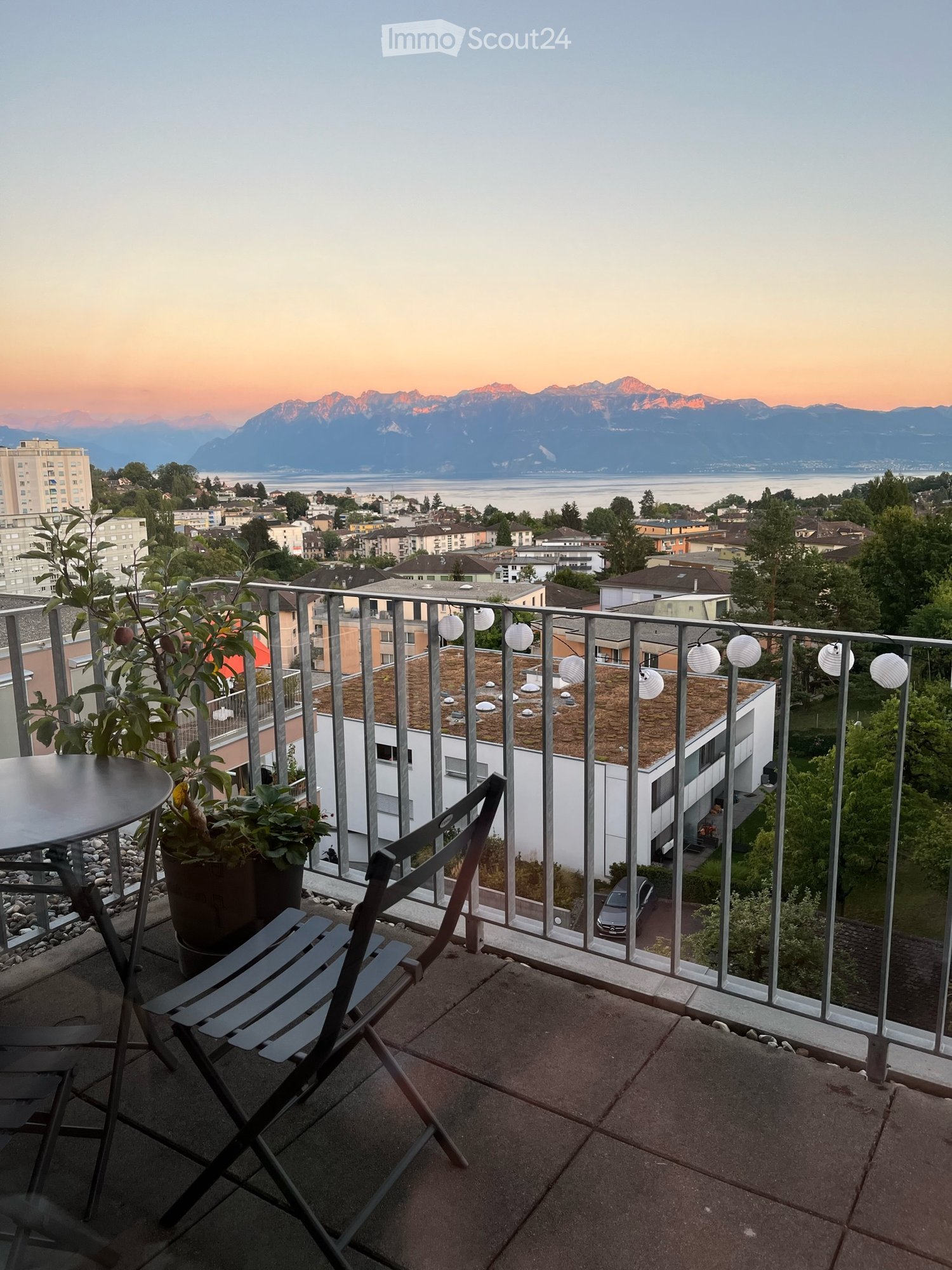 a balcony with metal railings, folding chairs, a small table, potted plants, an outdoor view of a city with mountains