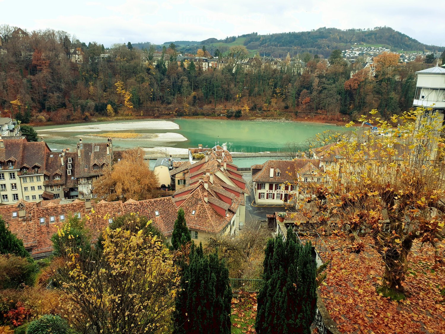 Aerial view of buildings with red roofs near a river, surrounded by trees and mountains