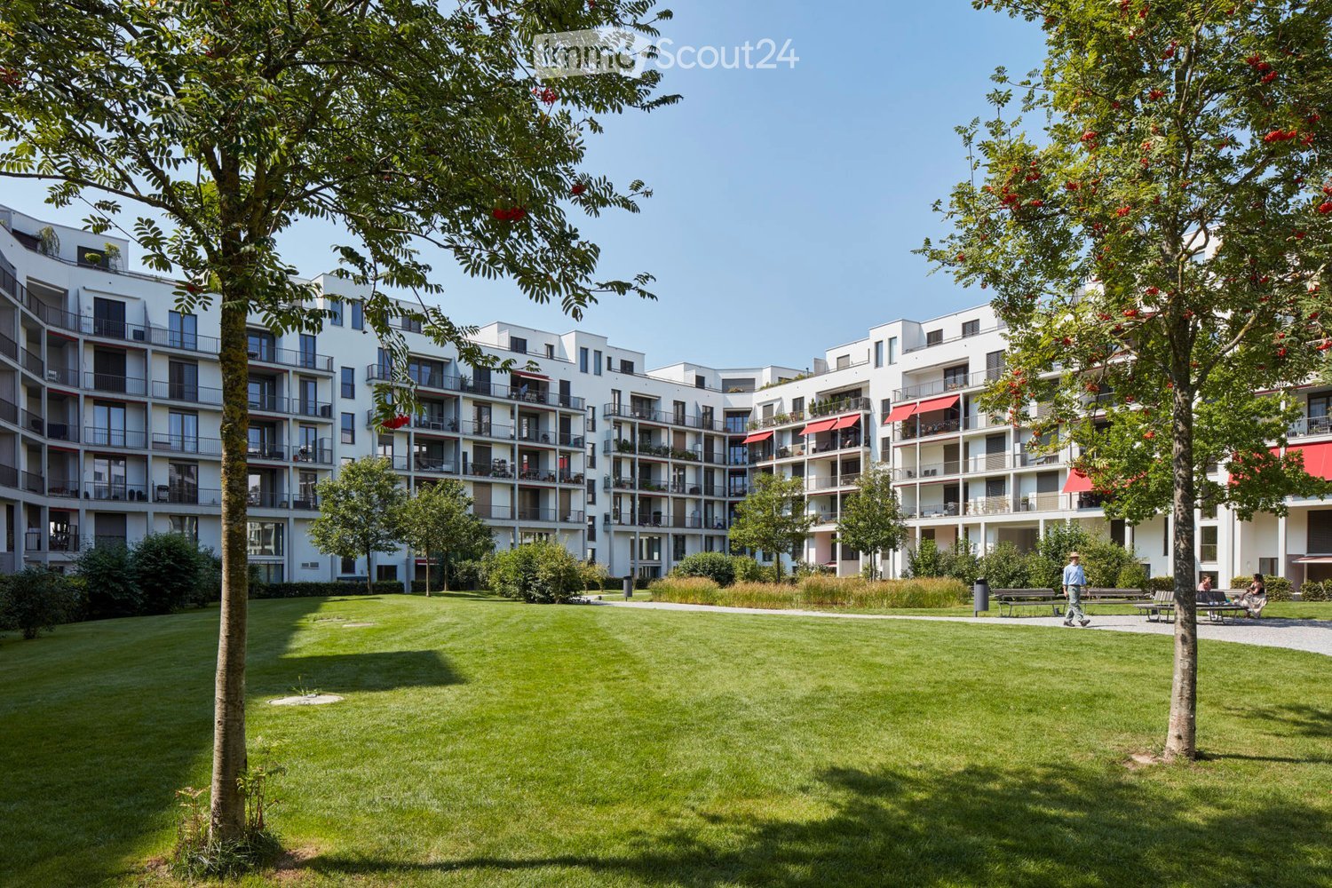 Modern multistory building with multiple balconies, landscaped lawn, trees, benches, walking paths