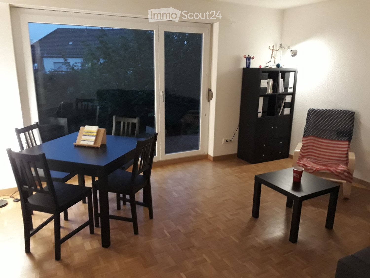 Dining area with black table and chairs, wooden floor, sliding door leading to a terrace with outdoor furniture, bookshelf, and armchair with blanket