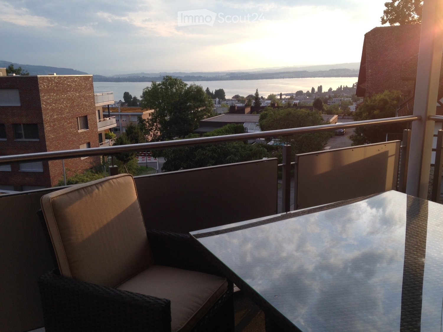 A balcony with a chair, table, and railings overlooking a landscape with a body of water, buildings, and trees