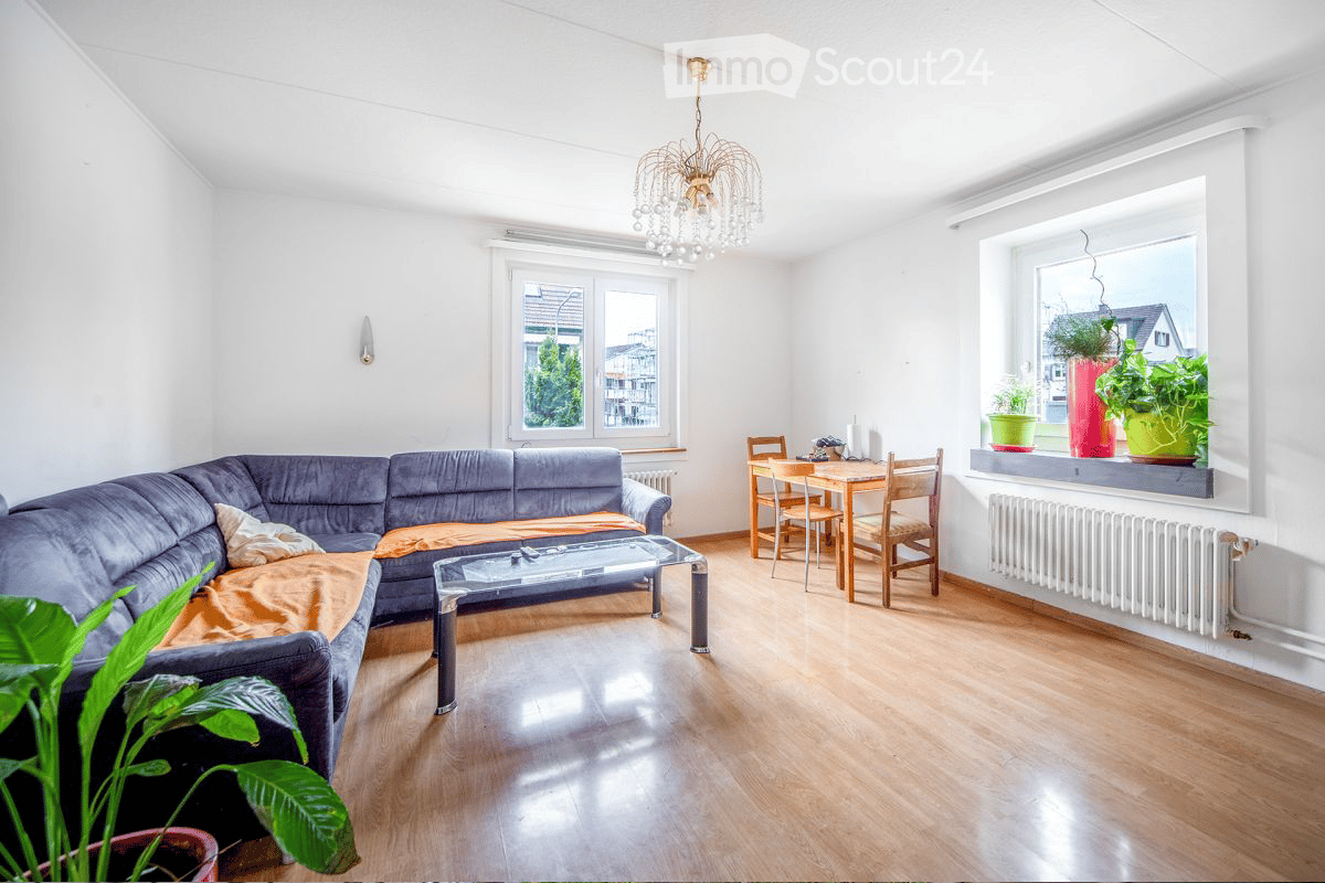 living room with sofa, coffee table, dining table, chairs, chandelier, white wall, wooden floor, window, radiator, plants on the window sill