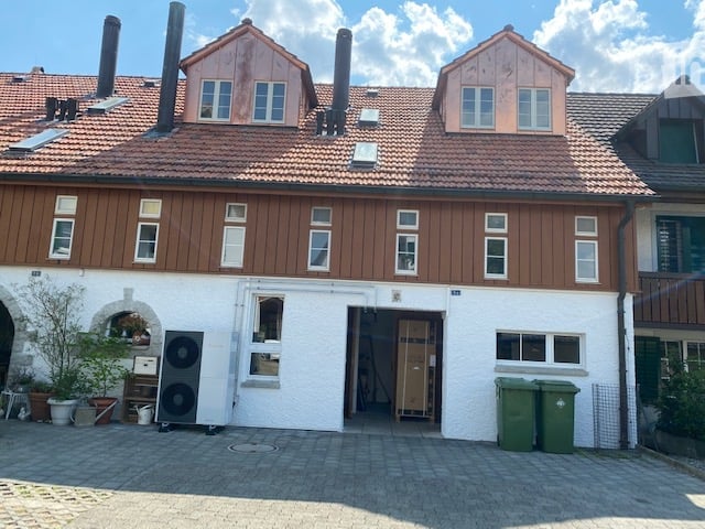 2-story building with red tile roof, wooden siding, and multiple windows. There is a paved parking area in front of the building.