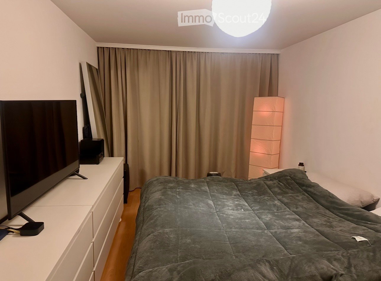 Bedroom with a bed, dresser with TV and speakers, beige curtains, floor lamp