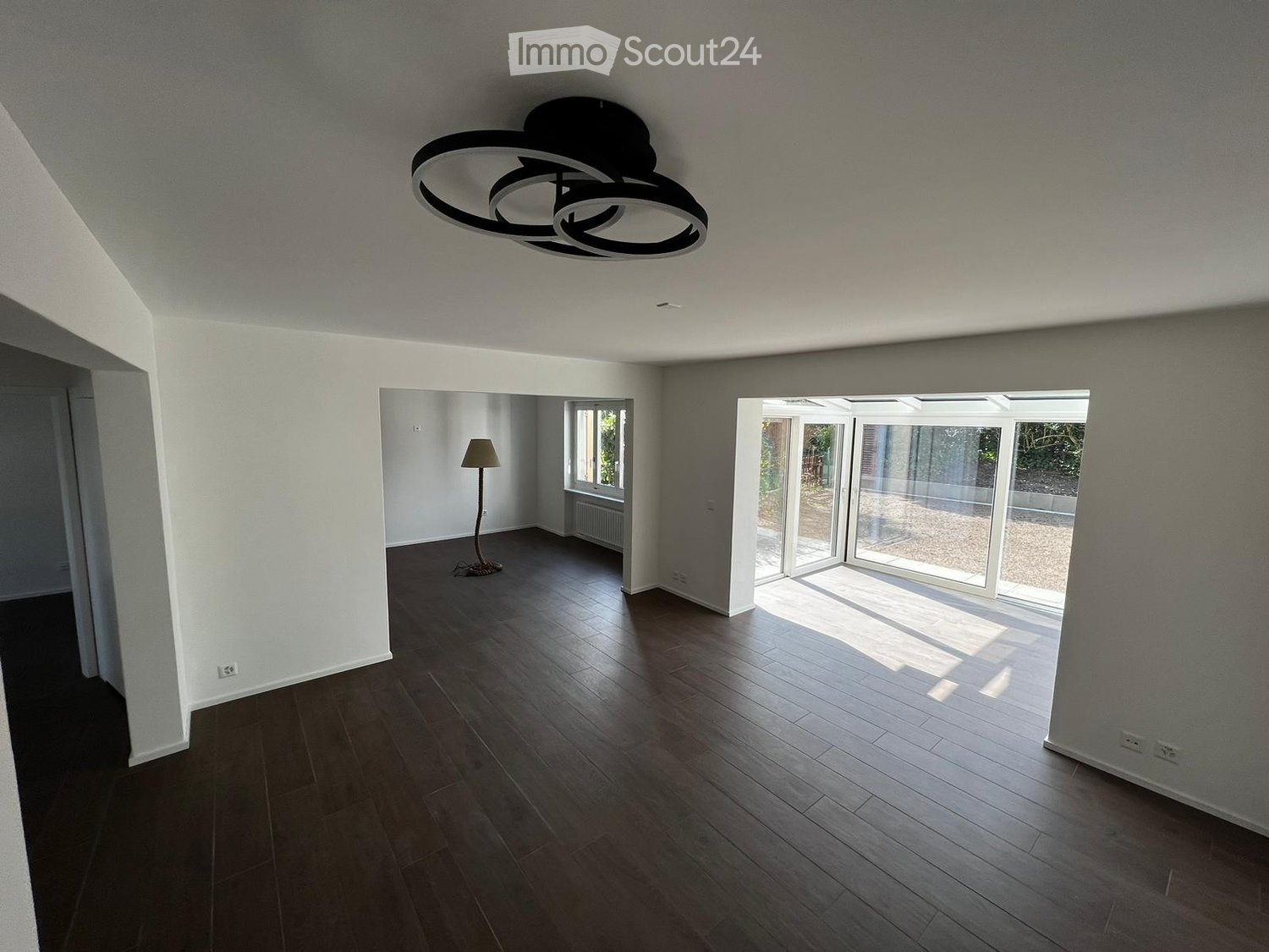 Empty room with white walls, wooden floor, ceiling lamp, large windows, glass doors leading to an outside area, power outlets