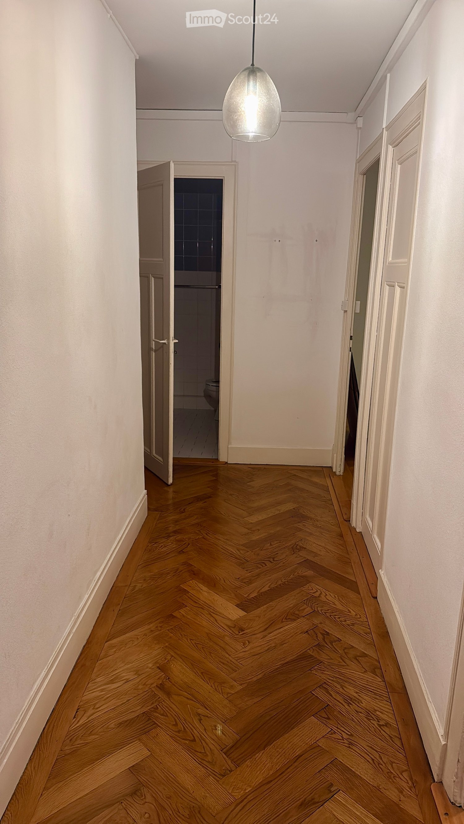 Empty hallway with wooden parquet floor and white walls, a door leading to a bathroom.