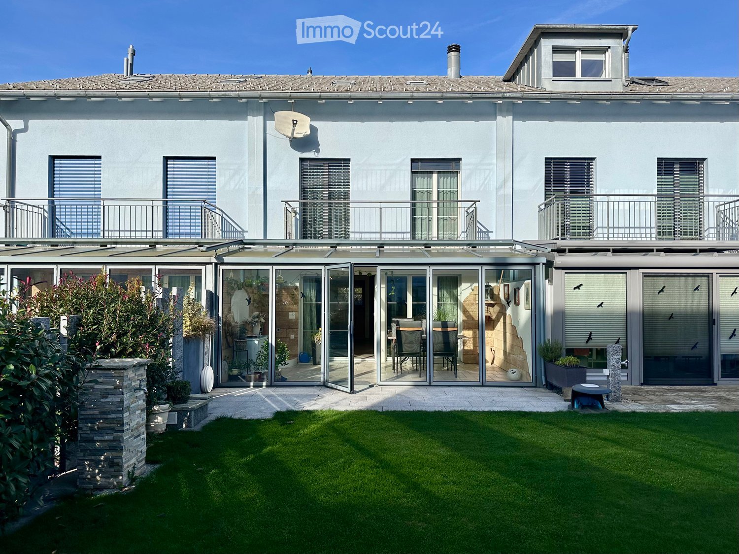 Detached house, gray exterior, double-glazed glass doors, glass balcony with metal railings, chimney, garden with plants and lawn