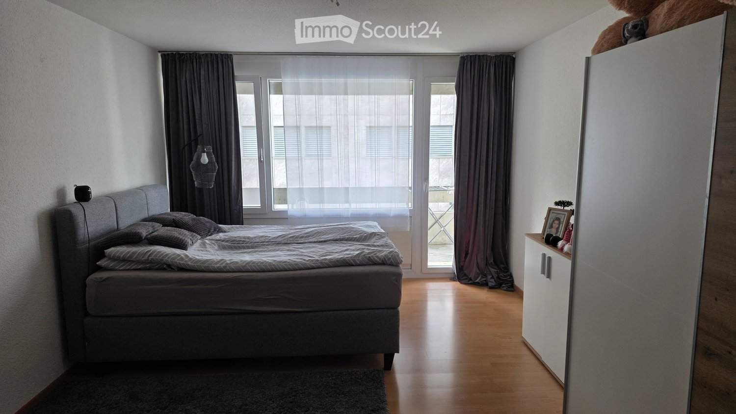 double bed, gray headboard, two pillows, window with curtains, wooden floor, closet