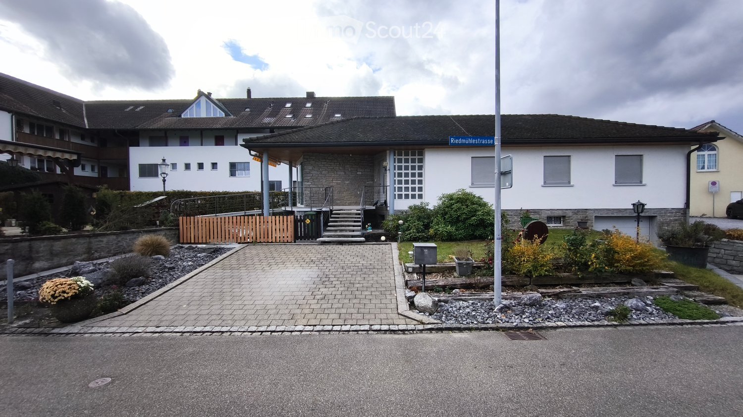 White house with stone walls, tiled roof, two large windows, stone steps leading up to the door, driveway, hedge, street sign