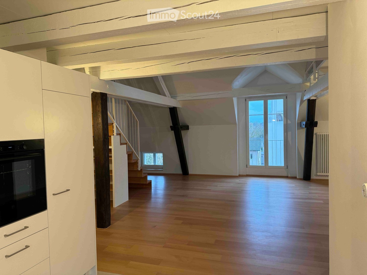 spacious room with wooden floor, white walls, large window, white cabinetry, oven, wooden stairs