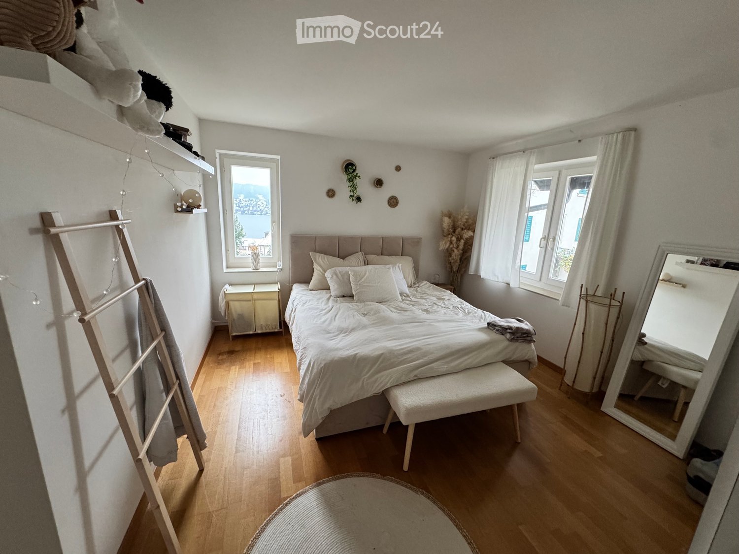 Large bed, white sheets, white headboard, white bench, white floor, white walls, two large windows, wooden ladder, rug, two white pillows, shelf with stuffed animals