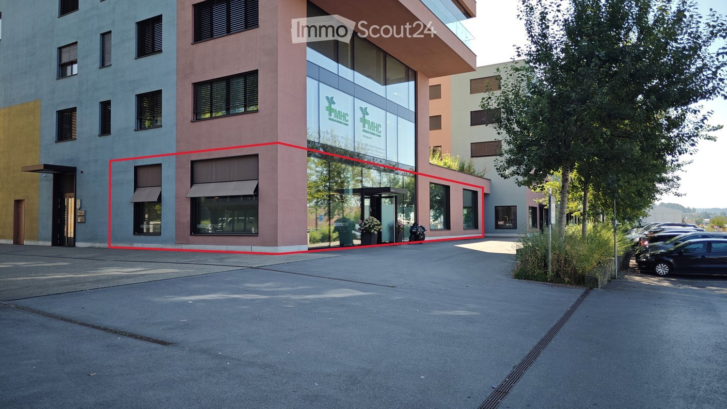 2 story building, glass windows, red marking, entrance, several windows, 2 flower pots, cars parked, several trees