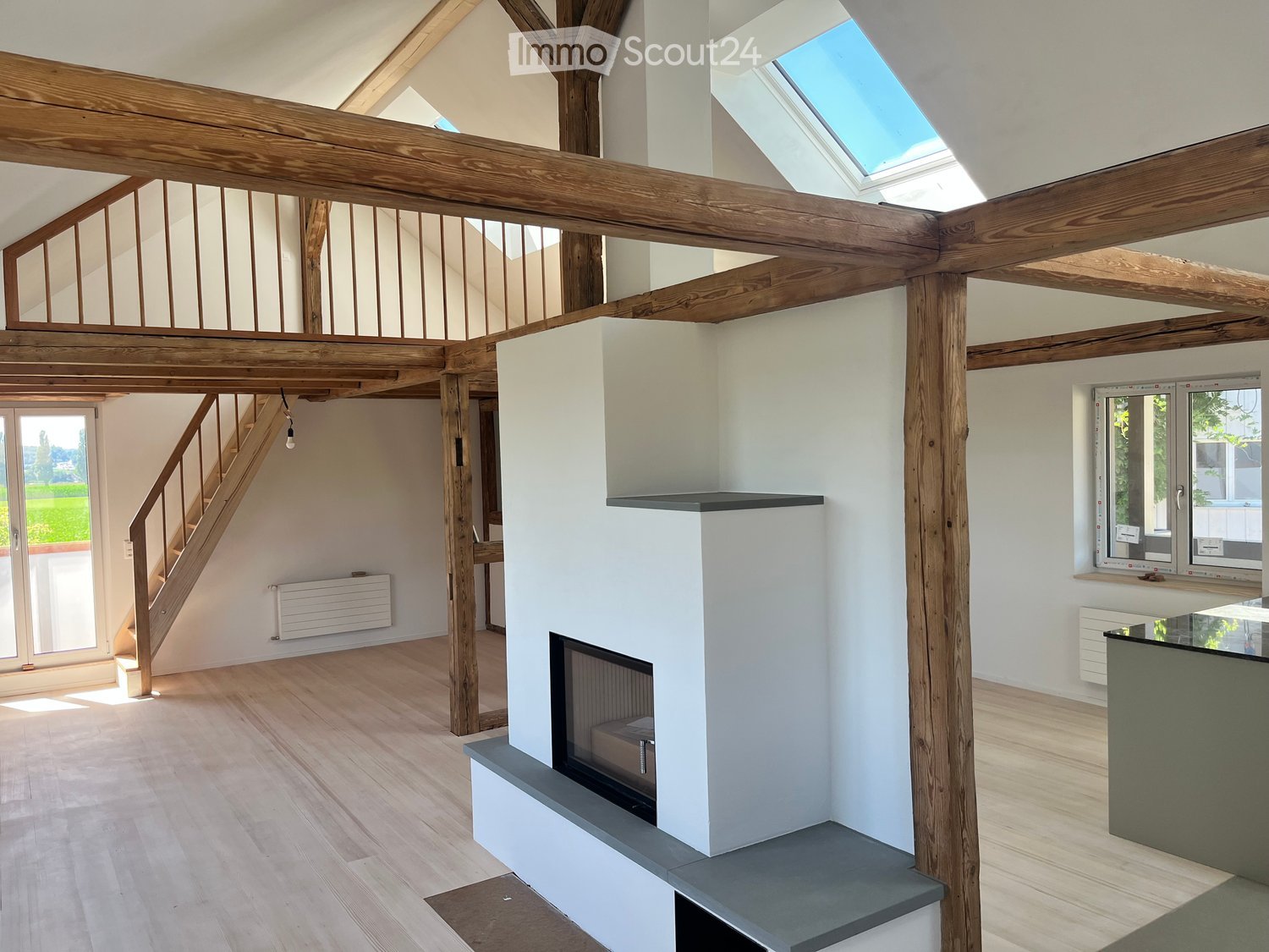 Open-concept living space with exposed wooden beams, a modern fireplace, and large windows providing natural light. The room has a minimalist and contemporary design with a neutral color palette.
