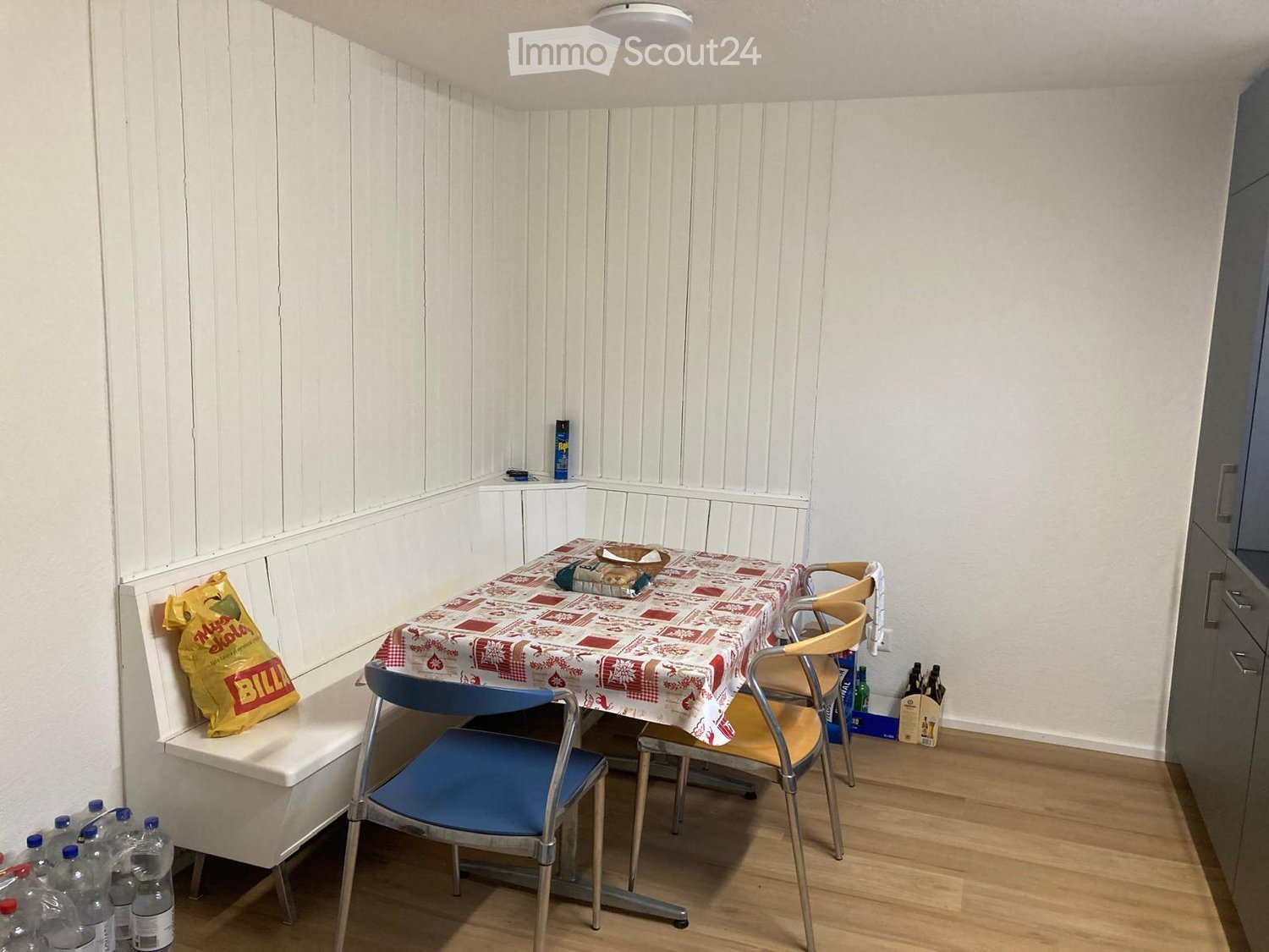 Dining area with a table, chairs, bench, white wooden walls, wooden floor, table with tablecloth, bags and bottles on floor.