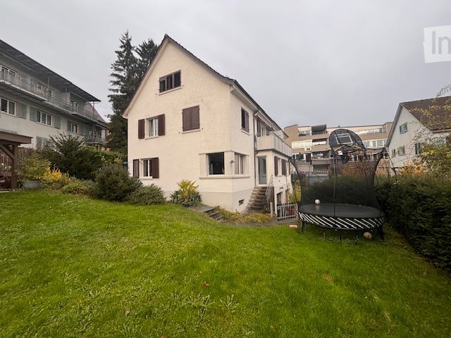 Large white house, front yard, trampoline, trees, bushes, staircase, balcony, green lawn