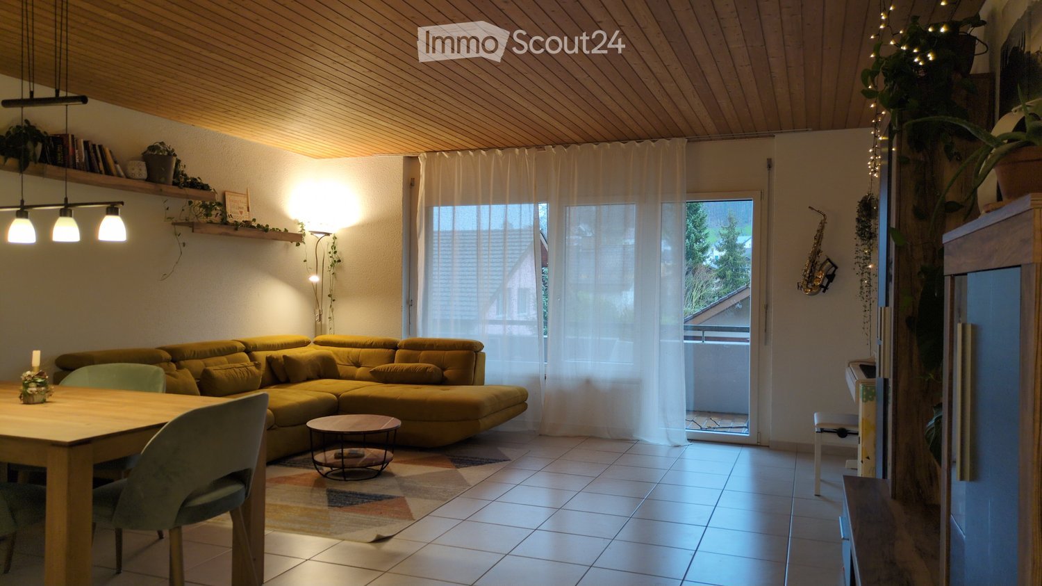 Modern open-plan living room with a large yellow couch, wooden table and chairs, a balcony with railings, carpet, wooden floor, white curtains, and floor-to-ceiling windows leading to a balcony.