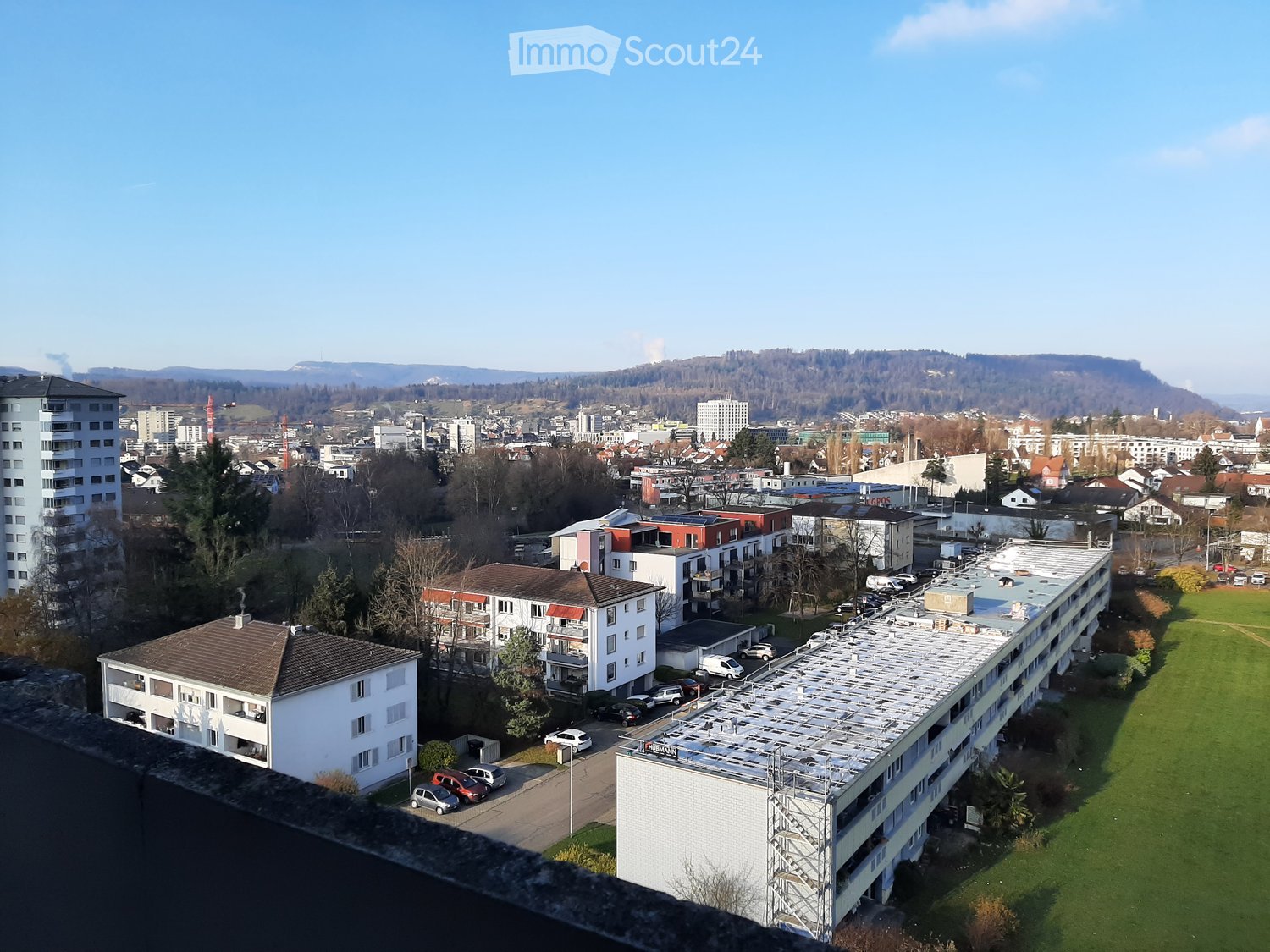 Cityscape with various residential buildings, a mix of modern and older architecture, surrounding greenery, a green field, distant mountains, and a clear blue sky.