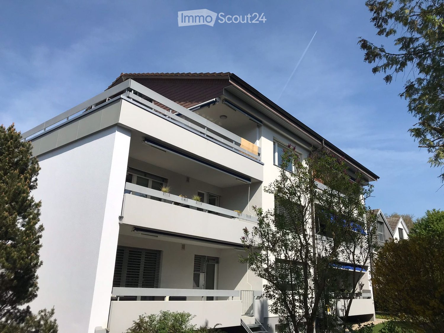 white apartment building, brown roof, 3 stories, balcony on each floor, surrounded by trees