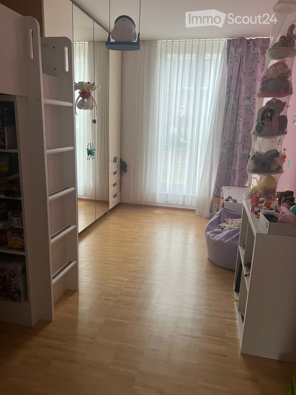 Children's room with wooden flooring, white shelves, a large mirror, a bean bag chair, and white curtains
