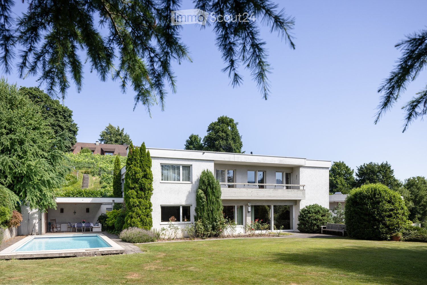 Modern white house, multiple windows, large front lawn, pool, trees, balcony, bench