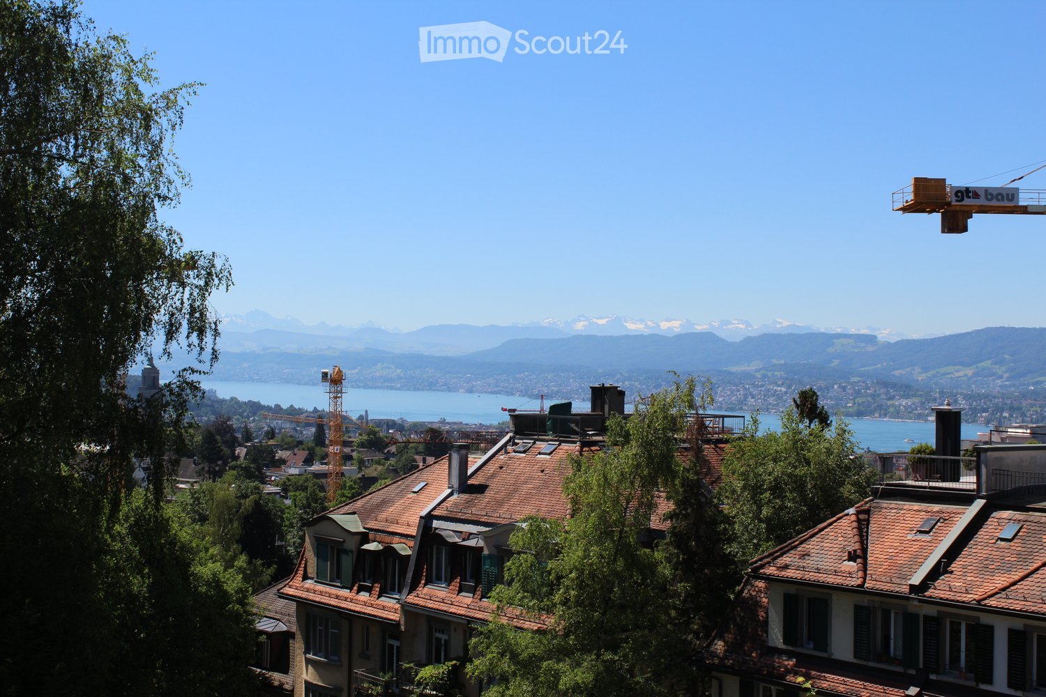 Aerial view of buildings with trees, a crane, lake, and mountains in the background