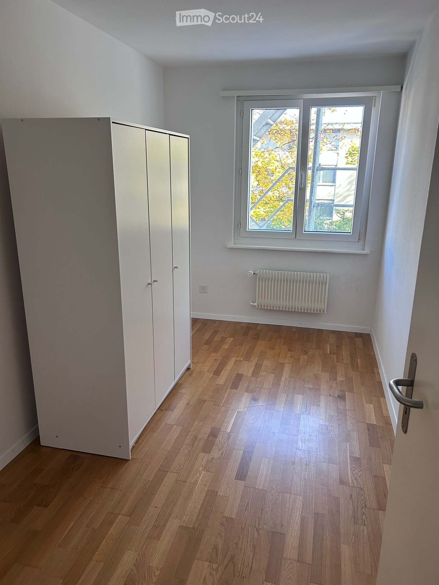 Empty room with wooden floors, white walls, large window, white wardrobe, and radiator