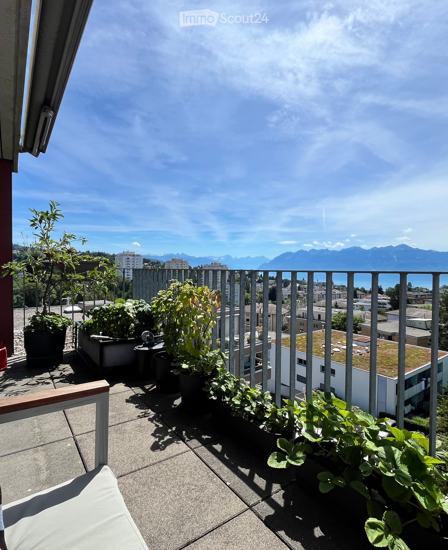 Terrace with metal railing, plants, trees, balcony chair, mountains in the background.