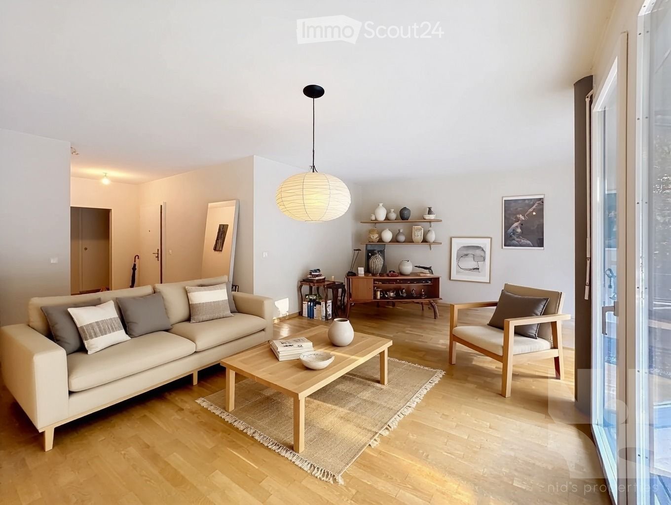 Modern living room with wooden floors, white walls, a chandelier, sofa, coffee table, bookshelves, picture frames, and a glass door leading to a balcony.