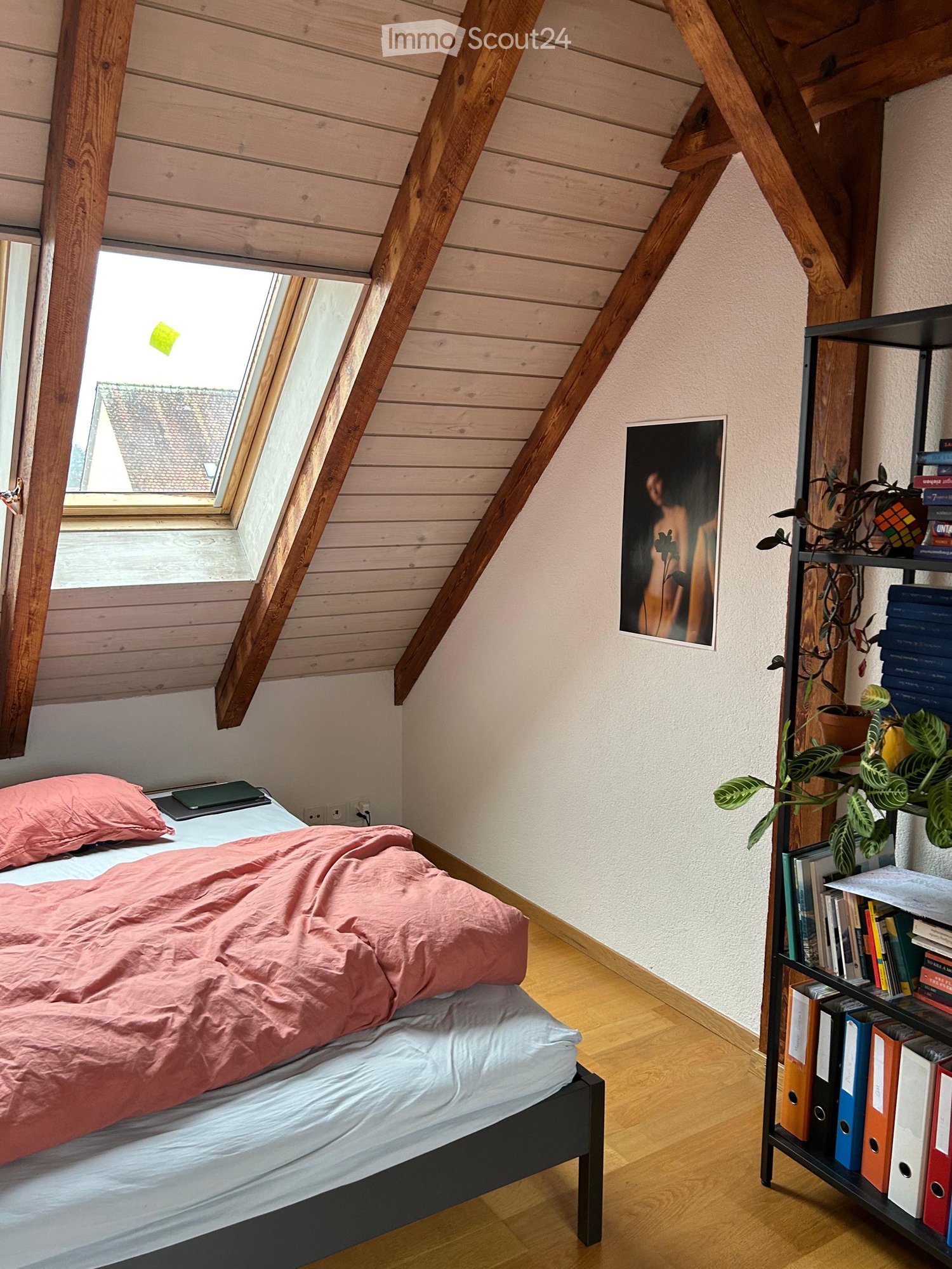 A small bedroom with wooden beams on the ceiling, a bed, a bedside table, a bookshelf with books and plants, and a skylight
