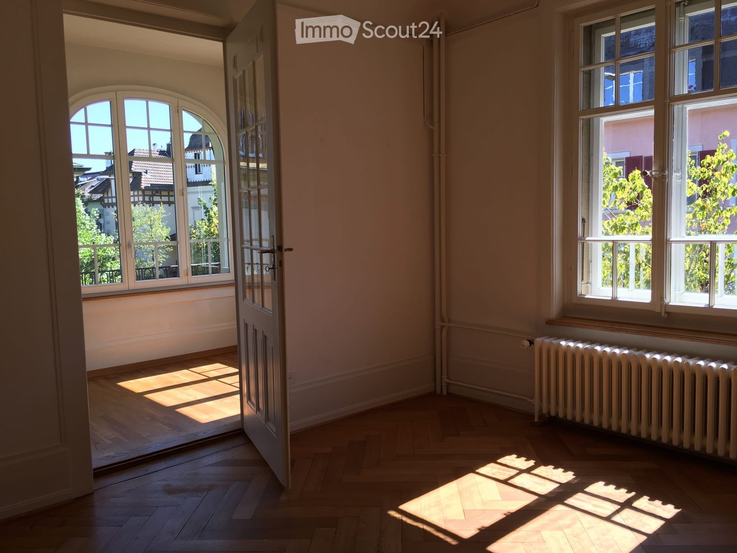 Empty room, large arched windows, wooden floors, radiator, open door to another room.