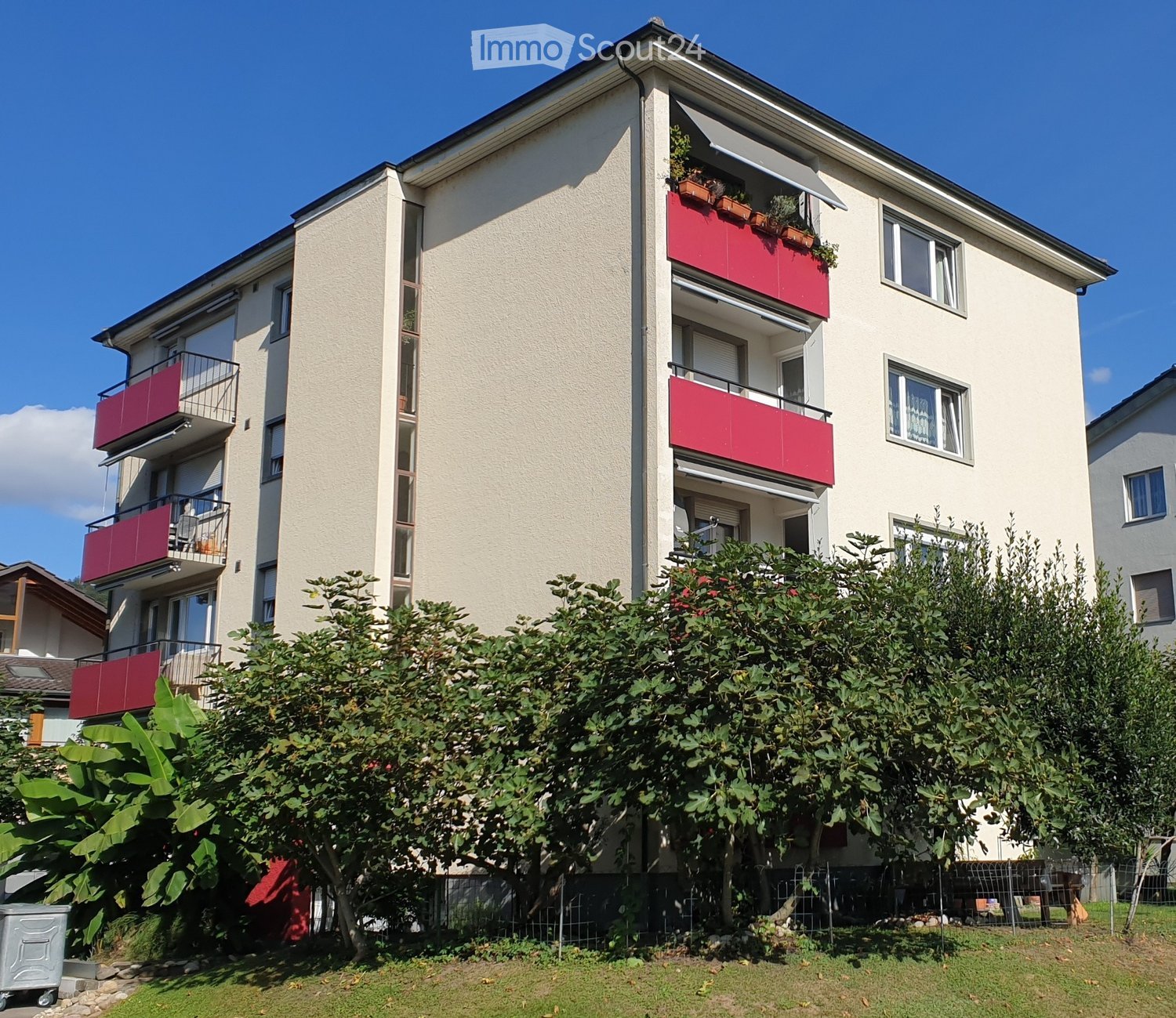 4-story apartment building with red balconies, white walls, multiple windows, shrubs, and bushes in front