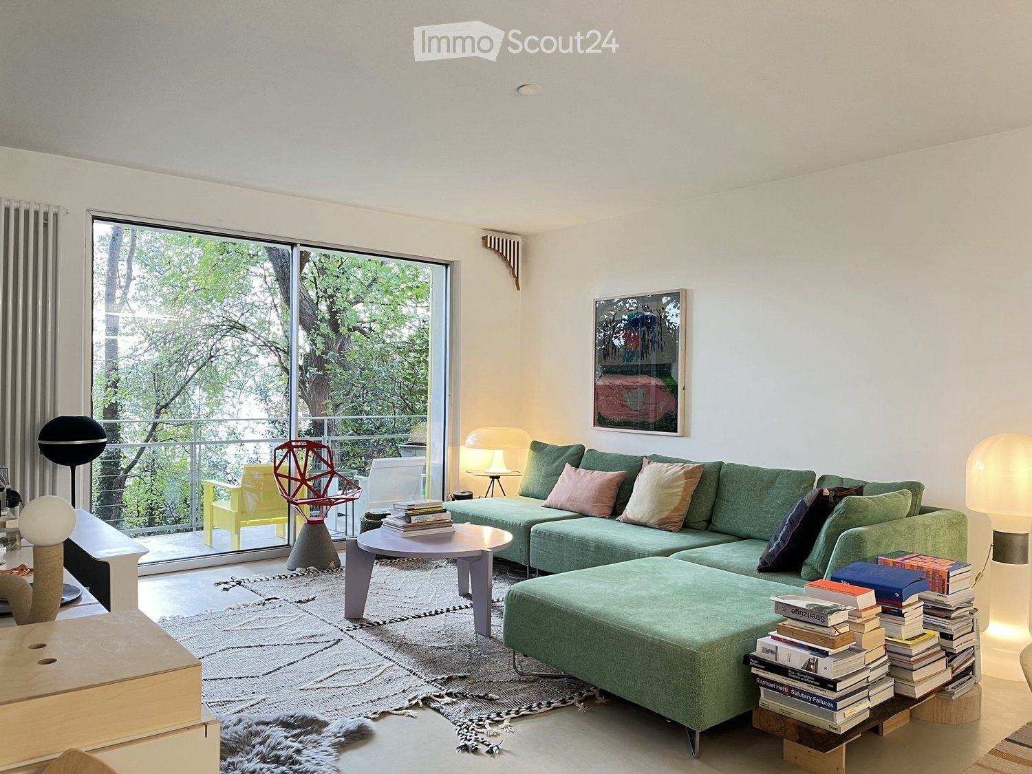 Green sofa, L shaped, large windows, balcony with tree view, large carpet, table with books, lamps